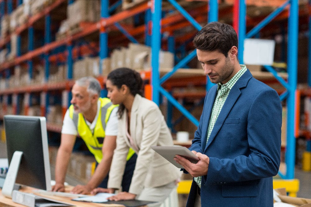 People standing in a warehouse with a man holding a tablet and two people in the background on a computer